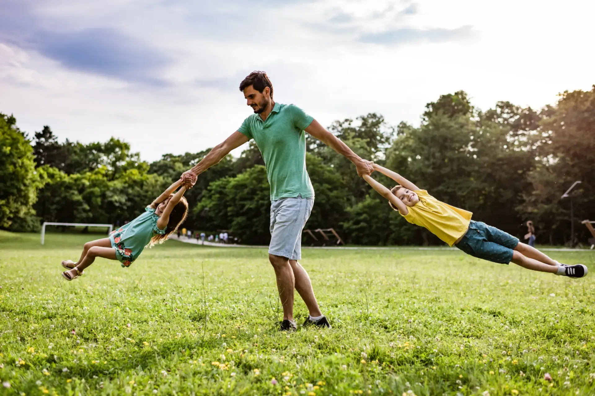 A father joyfully playing with his two children in a sunny park, surrounded by green grass and trees.